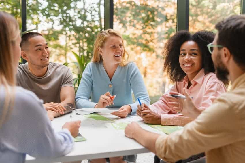 Groupe de collaborateurs en réunion de travail souriante autour d’une table, échangeant des idées et travaillant ensemble dans un environnement lumineux.
